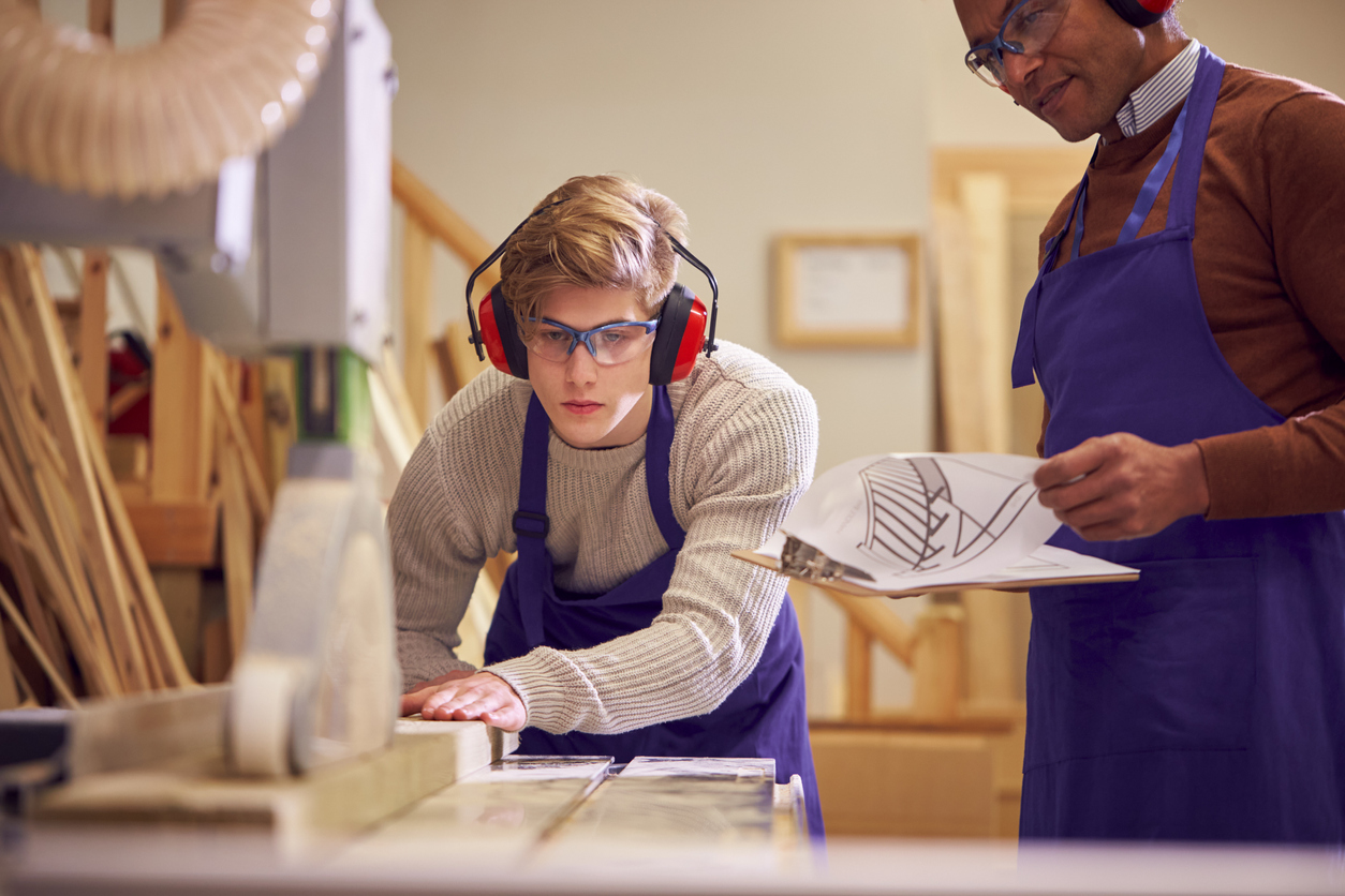 Tutor With Male Carpentry Student In Workshop Studying For Apprenticeship At College Using Bench Saw.