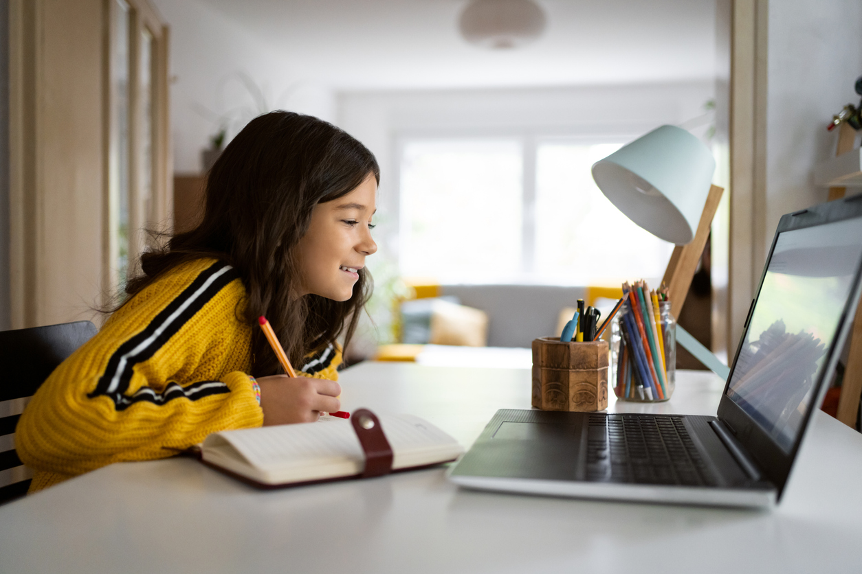 Stock young student in front of their laptop. They are sitting at a white table with a lamp and jars of pencils and writing in a notebook.