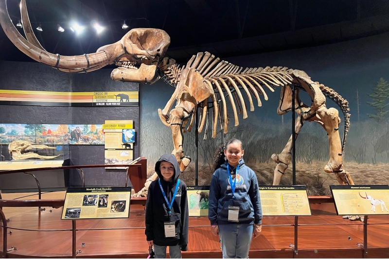 Two Insight PA students are standing in front of a skeleton of a wooly mammoth on a field trip to a museum.