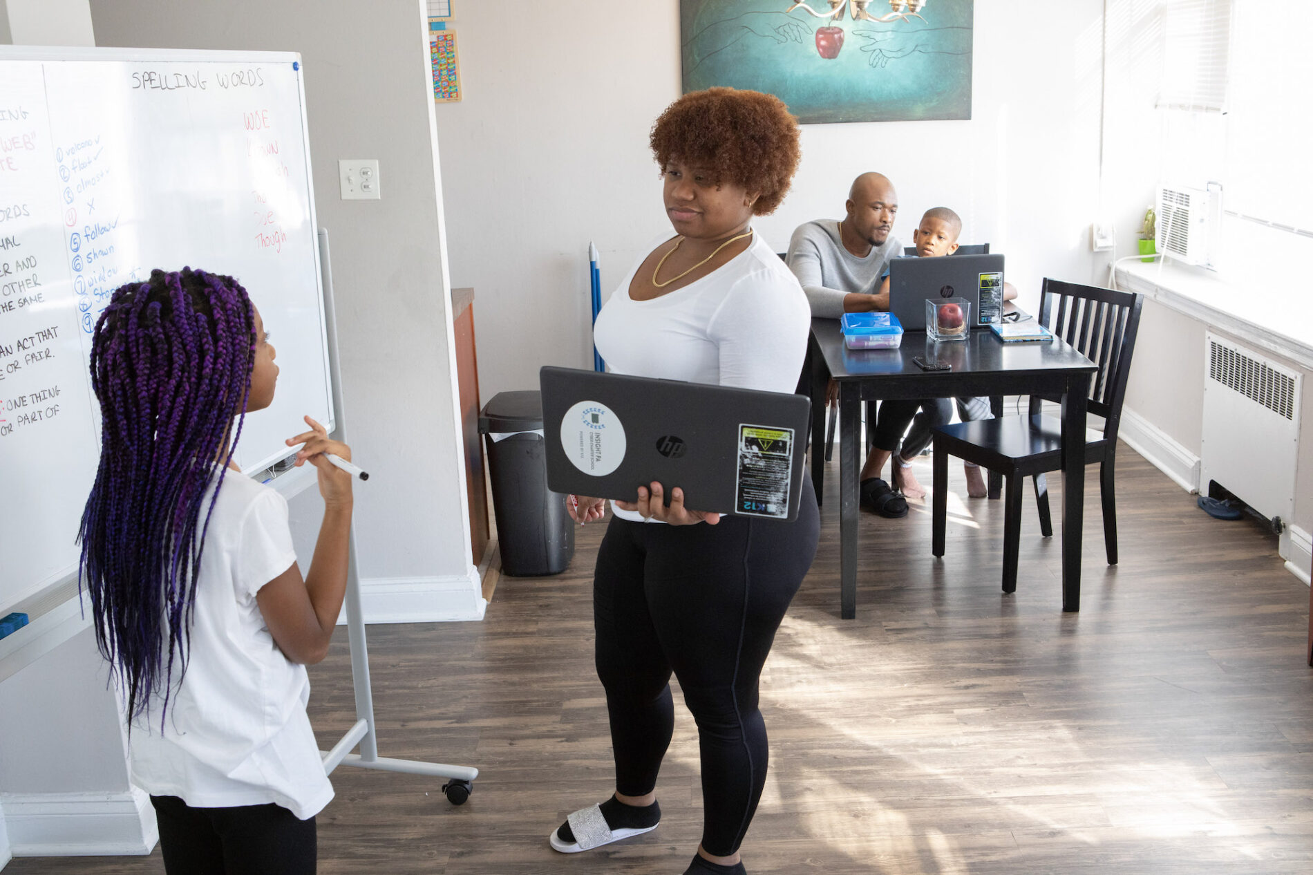 A mother and daughter work at a whiteboard and in the background a father and son are seated at a table