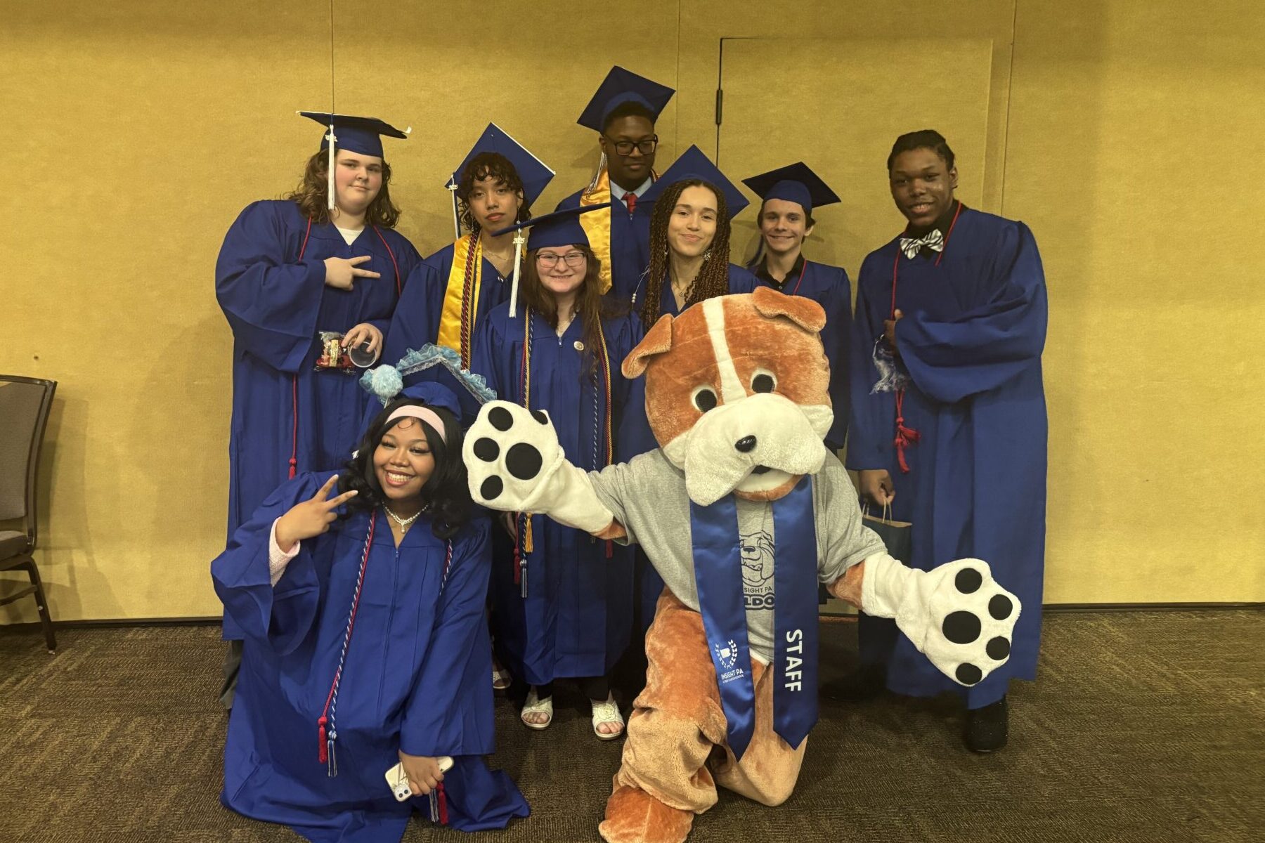 A group of Insight PA graduates is posing in their caps and gowns with the school’s bulldog mascot.