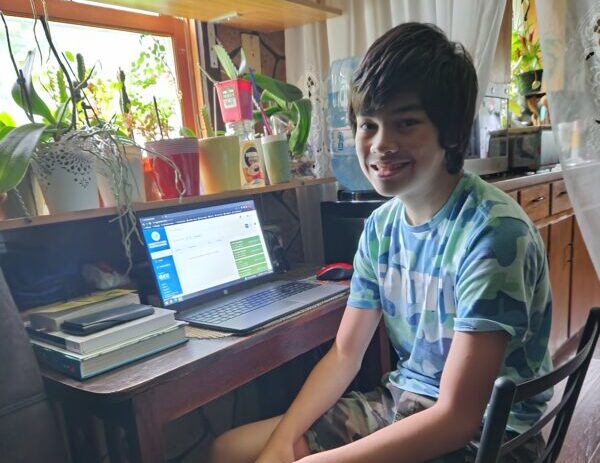 A smiling student sits at their desk with a laptop open. Colorful plants line the windowsill.