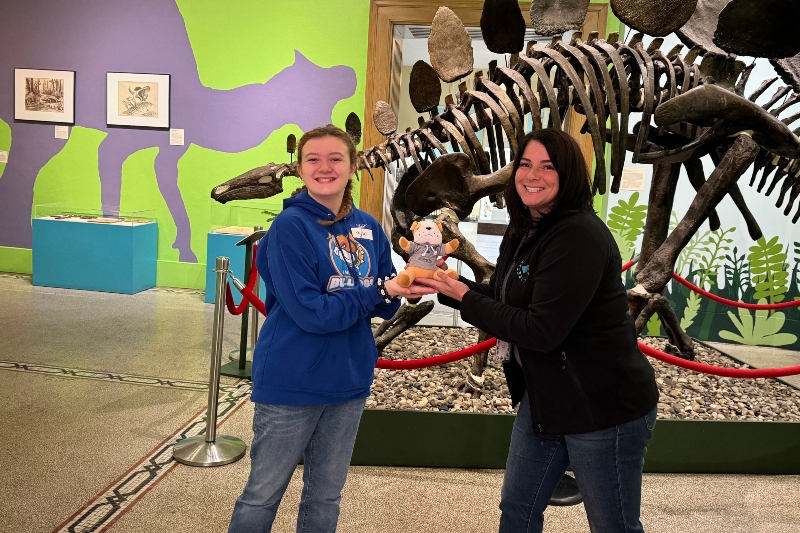 Community Engagement Coordinator Kristin Seidel poses with an Insight PA student holding a stuffed bulldog in front of a dinosaur skeleton at the Everhart Museum in Scranton, PA.